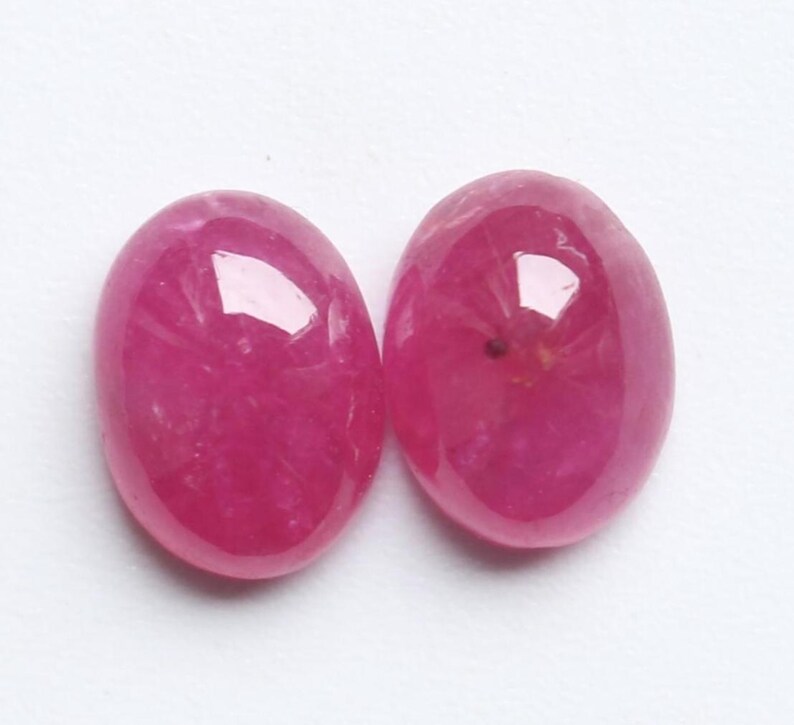 May include: Two oval-shaped, polished gemstones in a deep pink colour. The gems have a smooth, reflective surface, with subtle internal patterns. The gemstones are set against a plain white background.