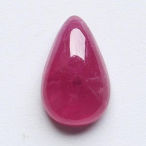 May include: A polished, teardrop-shaped ruby gemstone. The gem is a deep, saturated red colour with a smooth, reflective surface. The stone is set against a white background.