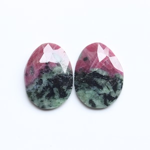 Może przedstawiać: Dwa owalne kamienie szlachetne o fasetowanej powierzchni. Kamienie prezentują mieszankę kolorów, w tym głęboką czerwień, czerń i zieleń. Kamienie szlachetne są umieszczone na białym tle.
