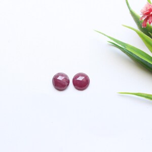 May include: Two round, faceted, deep red gemstones are displayed on a white surface. The gemstones are positioned near a sprig of green leaves and a small pink flower. The gemstones appear to be cabochons, possibly rubies, and are ready for jewelry making.