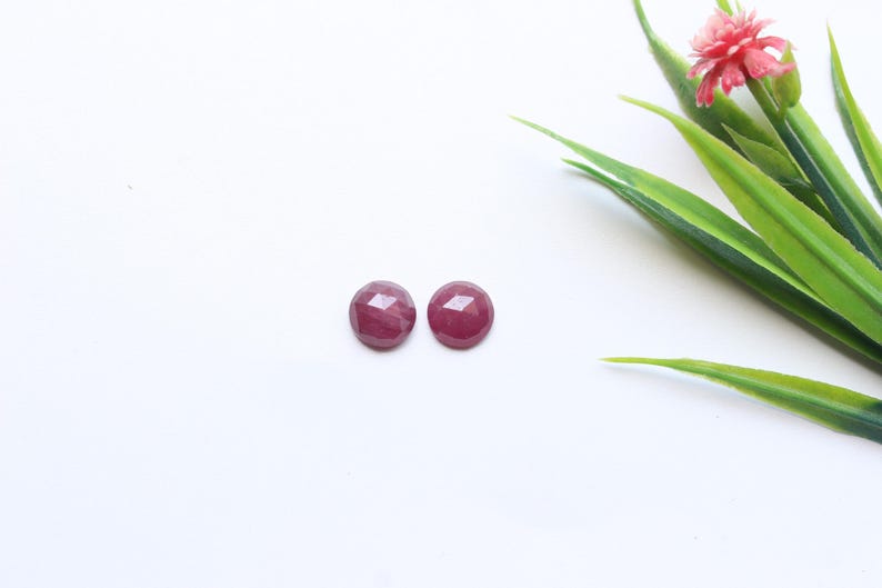 May include: Two round, faceted, deep red gemstones are displayed on a white surface. The gemstones are positioned near a sprig of green leaves and a small pink flower. The gemstones appear to be cabochons, possibly rubies, and are ready for jewelry making.