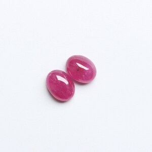 May include: Two oval-shaped, polished ruby gemstones. The gems are a vibrant, deep pinkish-red colour, with a smooth, reflective surface. They are set against a plain white background.