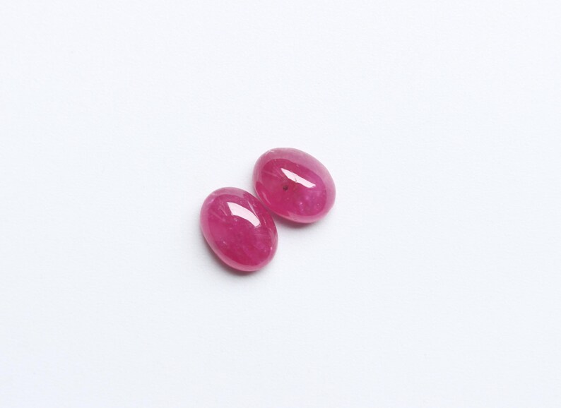 May include: Two oval-shaped, polished ruby gemstones. The gems are a vibrant, deep pinkish-red colour, with a smooth, reflective surface. They are set against a plain white background.