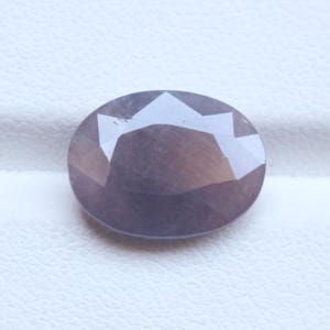 Op de afbeelding: Een gefacetteerde, ovaalvormige edelsteen met een rokerige, paarsbruine tint. De gepolijste oppervlakte heeft meerdere facetten die het licht reflecteren, waardoor een sprankelend effect ontstaat. De steen ligt tegen een witte achtergrond.