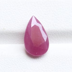 May include: A faceted, teardrop-shaped ruby gemstone. The gem is a deep, saturated pinkish-red color, with multiple facets that catch the light. The stone is set against a white background.