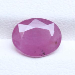 May include: An oval-shaped, faceted pink gemstone with a polished surface. The stone displays a vibrant magenta hue with subtle internal inclusions and geometric facets that reflect light across its translucent, rounded, and symmetrical body.