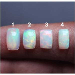 Pode incluir: Quatro opalas cabochão retangulares rotuladas 1, 2, 3 e 4. Cada pedra apresenta uma base branca leitosa com flashes iridescentes de luz verde, azul e rosa. As pedras estão dispostas em uma linha horizontal, exibindo uma superfície polida.