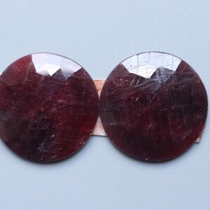 May include: Two round, faceted, dark red gemstones are displayed side-by-side. The gems have a polished, reflective surface with visible internal inclusions. They are mounted on a thin, light brown backing, set against a light blue-gray background.