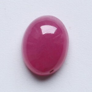 Könnte beinhalten: Ein oval geformter, polierter Edelstein in einem leuchtenden Pinkton. Die glatte, glänzende Oberfläche reflektiert das Licht. Der Stein liegt vor einem schlichten weißen Hintergrund, der seine Farbe und Form hervorhebt.