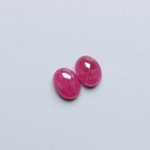 May include: Two oval-shaped, polished gemstones in a vibrant pink hue. The gems have a smooth, reflective surface, catching the light. The stones are set against a light grey background. These gems could be used for jewellery.