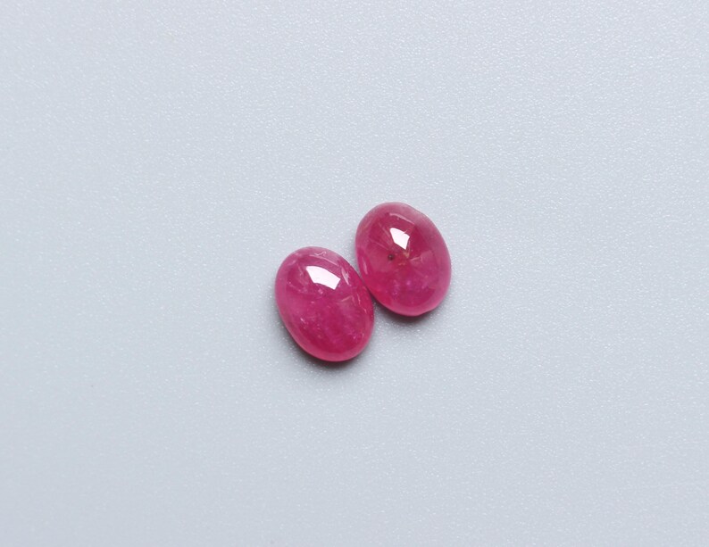 May include: Two oval-shaped, polished gemstones in a vibrant pink hue. The gems have a smooth, reflective surface, catching the light. The stones are set against a light grey background. These gems could be used for jewellery.
