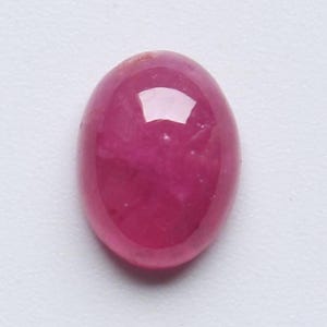 Könnte beinhalten: Ein polierter, oval geformter Rubin-Edelstein. Der Edelstein hat eine tiefe, gesättigte, rötlich-rosa Farbe mit einer glatten, reflektierenden Oberfläche. Der Stein liegt vor einem schlichten, weißen Hintergrund.