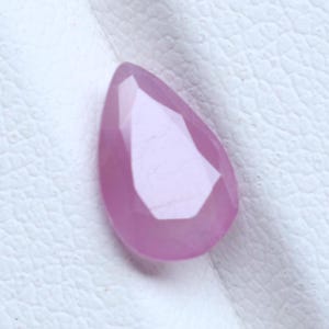 May include: A pear-shaped, faceted gemstone with a translucent light pink color. The stone features a pointed top and a rounded base, with multiple flat, geometric surfaces reflecting light across its polished, semi-transparent surface.