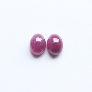 May include: Two oval-shaped, polished gemstones in a deep magenta hue are presented against a white backdrop. The gems have a smooth, glossy surface, reflecting light. These gemstones are likely rubies, ideal for jewelry.