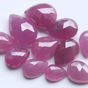 Puede incluir: Una colección de gemas facetadas, en forma de lágrima y ovaladas, en varios tonos de rosa y rojo. Las gemas están dispuestas sobre una superficie blanca, mostrando sus superficies pulidas y reflejos internos. Las gemas son probablemente rubíes.