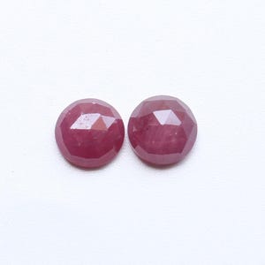 Puede incluir: Dos gemas redondas de rubí facetadas. Los rubíes son de color rojo intenso y tienen una superficie pulida y reflectante. Las gemas están sobre un fondo blanco.