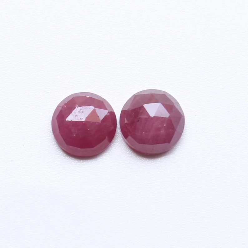 May include: Two round, faceted ruby gemstones. The rubies are a deep red color and have a polished, reflective surface. The gemstones are set against a white background.