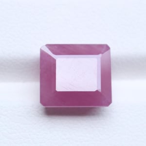 Peut inclure: Une pierre précieuse de taille carrée avec une teinte rose vive et une surface à facettes. La pierre présente une table plate et des bords inclinés qui reflètent la lumière, créant un aspect géométrique avec une finition lisse et polie.