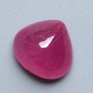 May include: A smooth, polished, cabochon-cut ruby gemstone. The gem is a deep, saturated pinkish-red color with a high luster, reflecting light. The shape is rounded and irregular, with a slightly flattened back.