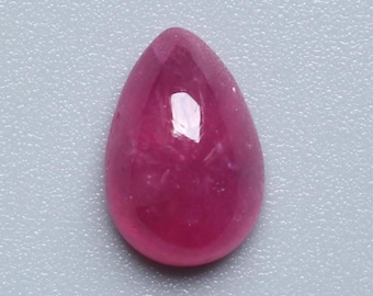 Puede incluir: Una gema de rubí pulido en forma de lágrima. La gema es de un color rojo rosado profundo y saturado con una superficie lisa y reflectante. La piedra está sobre un fondo gris claro.