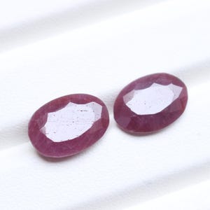 May include: Two oval-shaped, faceted rubies with a deep magenta-red hue. Each gemstone features a polished surface with multiple geometric facets that reflect light, showcasing a translucent quality and a smooth, rounded edge profile.