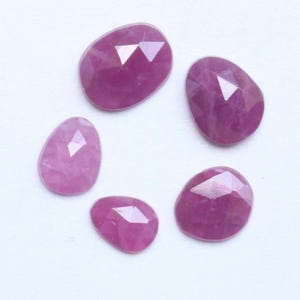 Pode incluir: Cinco pedras preciosas de rubi facetadas, em forma oval e de tamanhos variados. As gemas são de cor vermelho-rosada profunda e têm uma superfície polida e reflexiva. As pedras estão dispostas sobre um fundo branco.