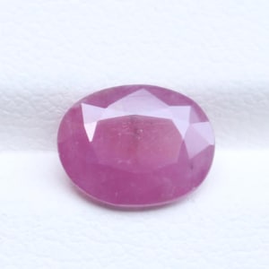 May include: A vibrant pink gemstone with an oval shape and a faceted cut. The stone features a polished surface with multiple triangular and trapezoidal facets that reflect light, creating a clear, symmetrical pattern across its top surface.