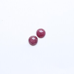 May include: Two round, faceted, deep red gemstones are displayed against a white background. The gems have a polished, reflective surface, and their circular shape is emphasized by the multiple facets. These gemstones are suitable for jewelry making.