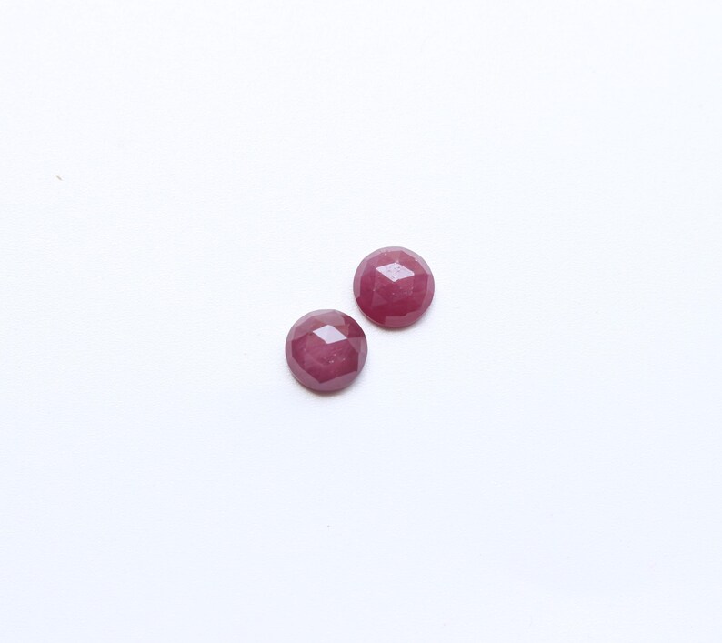 May include: Two round, faceted, deep red gemstones are displayed against a white background. The gems have a polished, reflective surface, and their circular shape is emphasized by the multiple facets. These gemstones are suitable for jewelry making.