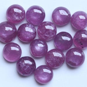 Puede incluir: Una colección de gemas redondas y pulidas en tonos rosados y púrpuras. Las gemas tienen una superficie lisa y reflectante, con inclusiones internas visibles. Las piedras están dispuestas juntas, mostrando su color y textura.