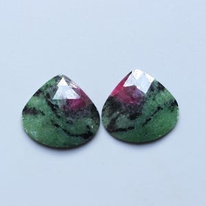 May include: Two teardrop-shaped gemstones with a faceted top. The gems feature a mix of green, black, and red hues. The stones are set against a light gray background.
