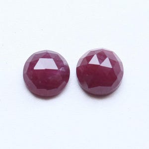 May include: Two round, faceted, deep red gemstones. The gems have a polished, reflective surface and are set against a white background. The gemstones are likely rubies, suitable for jewelry making.