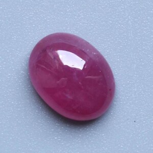 Könnte beinhalten: Ein polierter, ovaler Rubin-Edelstein. Der Edelstein hat eine tiefe, gesättigte, rötlich-rosa Farbe mit glänzender Oberfläche. Der Stein reflektiert Licht und erzeugt Glanzlichter. Der Rubin liegt vor einem hellblau-grauen Hintergrund.