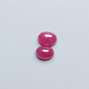 May include: Two oval-shaped, polished ruby gemstones. The rubies are a vibrant, deep pink-red colour, with a smooth, reflective surface. The gemstones are displayed against a light grey background.