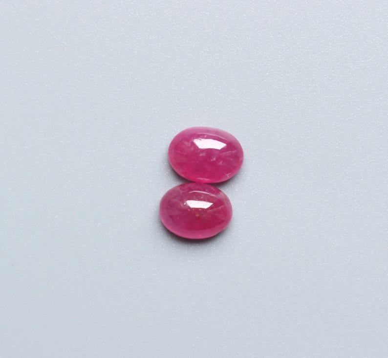 May include: Two oval-shaped, polished ruby gemstones. The rubies are a vibrant, deep pink-red colour, with a smooth, reflective surface. The gemstones are displayed against a light grey background.