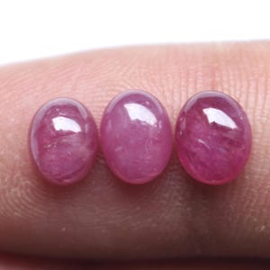May include: Three oval-shaped, cabochon-cut gemstones with a vibrant, translucent magenta-pink color. Each stone features a smooth, polished, domed surface that reflects light, showcasing internal textures and natural variations within the material.
