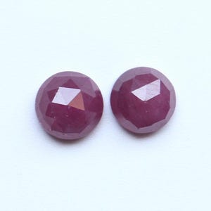 Puede incluir: Dos gemas facetadas de forma ovalada con un tono rojo intenso. Las gemas tienen una superficie pulida y reflectante, mostrando múltiples facetas que capturan la luz. Probablemente son rubíes, a menudo utilizados en joyería.