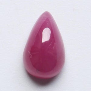 May include: A polished, teardrop-shaped gemstone in a deep, vibrant magenta hue. The gem has a smooth, reflective surface, with a subtle internal glow. The stone's form is elongated, tapering to a rounded point.
