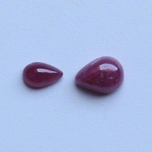 May include: Two teardrop-shaped, polished gemstones in varying sizes. The gems are a deep, rich red color with a glossy finish, reflecting light. The background is a plain, light blue.