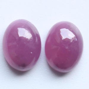 Puede incluir: Dos gemas ovaladas y pulidas en un tono rosa intenso. Las piedras tienen una superficie lisa y brillante, que refleja la luz. Las gemas están sobre un fondo blanco. Ideales para la creación de joyas.