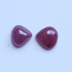 May include: Two polished, oval-shaped gemstones with a deep pinkish-red hue. The stones have a smooth, glossy surface and are set against a white background. These gemstones are likely rubies, perfect for jewelry making.