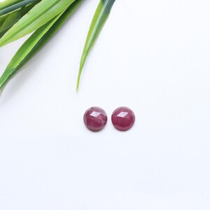 May include: Two round, faceted, deep red gemstones are displayed on a white surface. The gemstones are positioned near green leaves and a small red flower. These gemstones could be used for jewelry making.