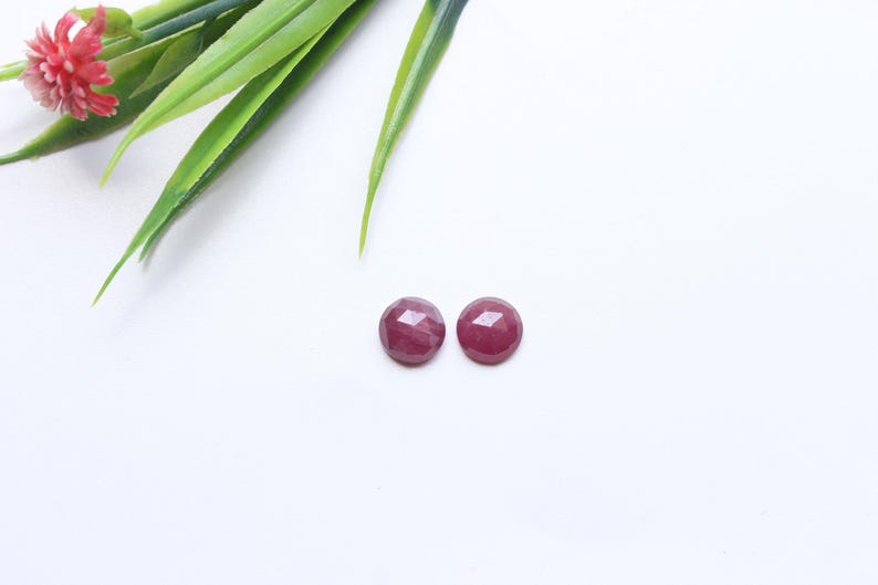 May include: Two round, faceted, deep red gemstones are displayed on a white surface. The gemstones are positioned near green leaves and a small red flower. These gemstones could be used for jewelry making.