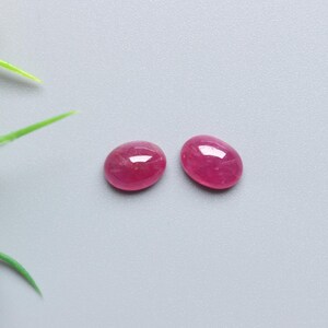 May include: Two oval-shaped, polished ruby gemstones with a vibrant red hue. The gemstones are displayed on a grey surface, with artificial greenery and yellow and purple flowers in the background.