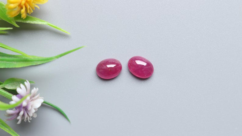 May include: Two oval-shaped, polished ruby gemstones with a vibrant red hue. The gemstones are displayed on a grey surface, with artificial greenery and yellow and purple flowers in the background.