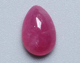 Puede incluir: Una gema de rubí pulida en forma de lágrima. La gema es de un color rojo rosado intenso y saturado, con una superficie lisa y reflectante. La piedra se encuentra sobre un fondo gris claro.