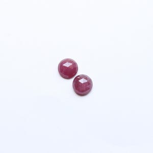 May include: Two faceted, round, deep red gemstones are displayed against a white background. The gems have a polished, reflective surface, and their shape is emphasized by the multiple facets. These gemstones could be used in jewelry.