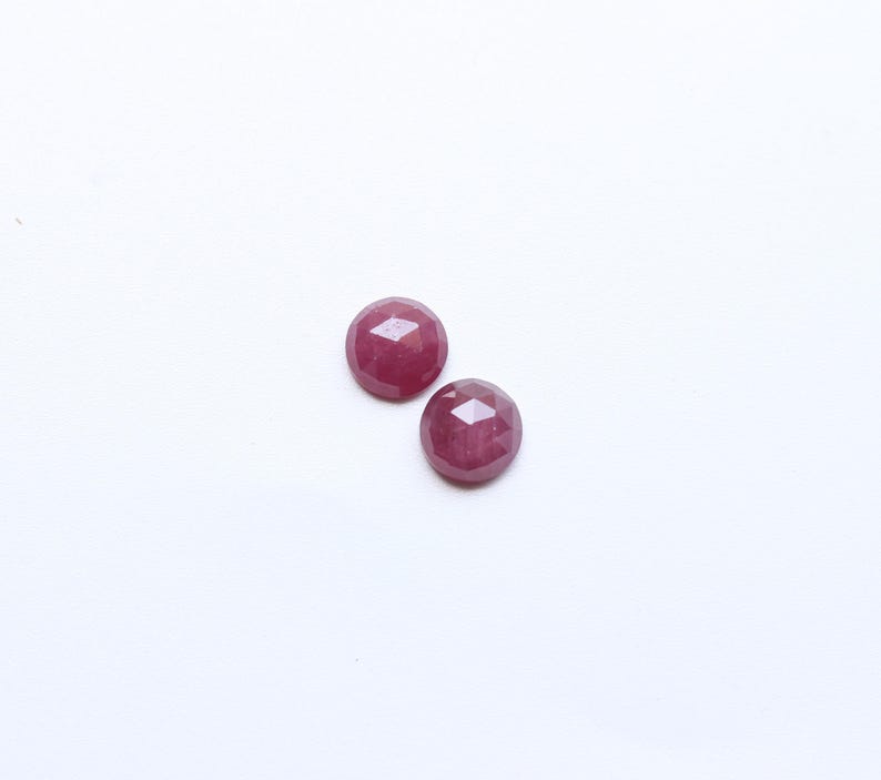 May include: Two faceted, round, deep red gemstones are displayed against a white background. The gems have a polished, reflective surface, and their shape is emphasized by the multiple facets. These gemstones could be used in jewelry.