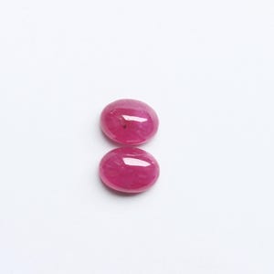 May include: Two oval-shaped, polished ruby gemstones. The rubies are a vibrant, deep pinkish-red colour, with a smooth, reflective surface. They are set against a plain white background, highlighting their colour and shape.