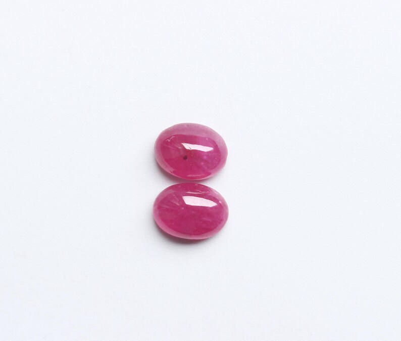 May include: Two oval-shaped, polished ruby gemstones. The rubies are a vibrant, deep pinkish-red colour, with a smooth, reflective surface. They are set against a plain white background, highlighting their colour and shape.